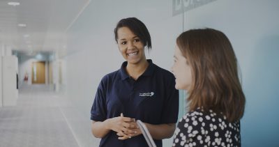 Two female co-workers standing against a wall in an office corridor having a conversation and smiling