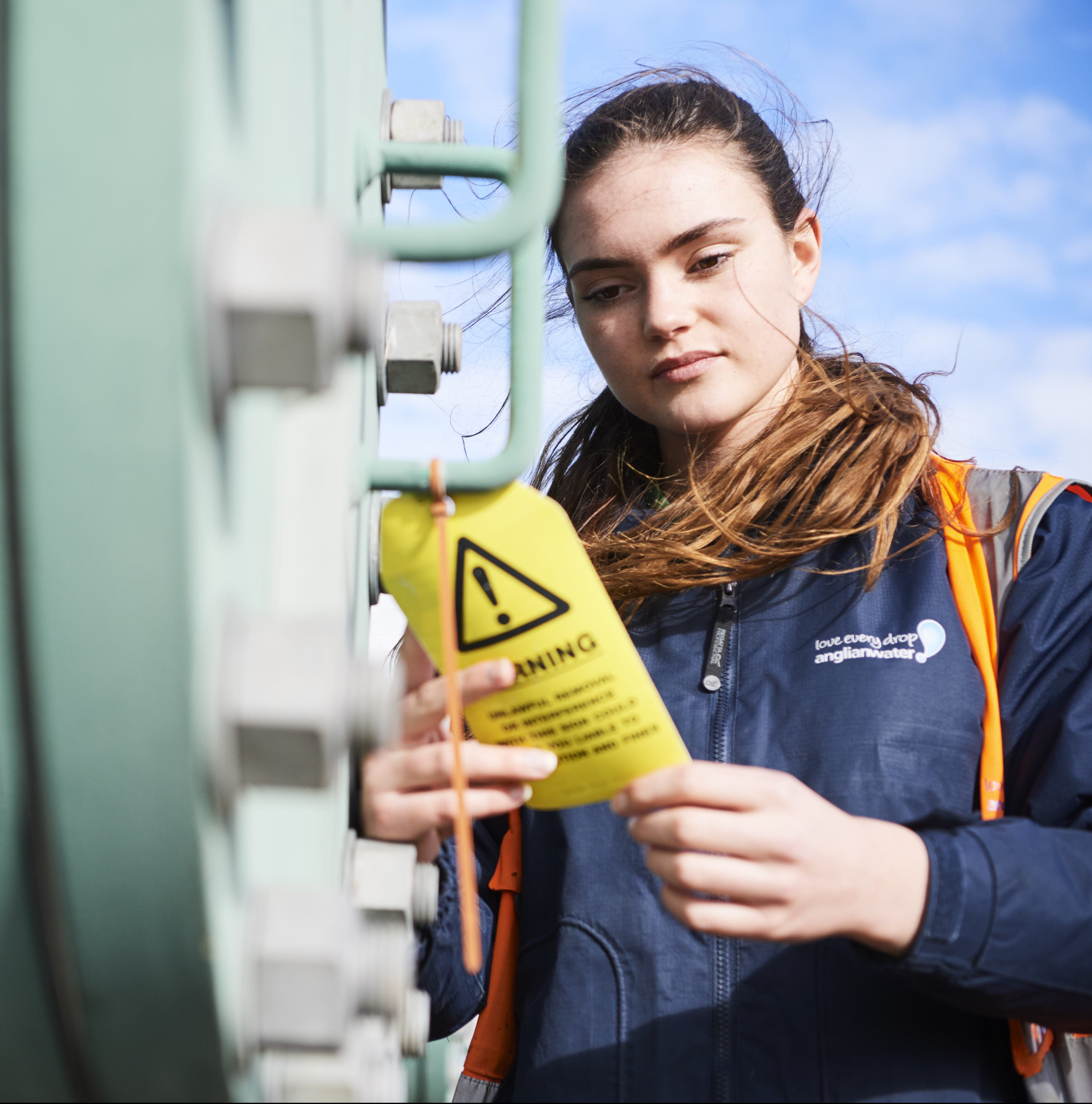 Close up from below shot of female apprentice in high vis jacket looking at yellow warning label on machine outside