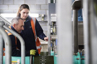 2 colleagues at a treatment facility, one male and one female, in high vis vests looking at a panel of instruments