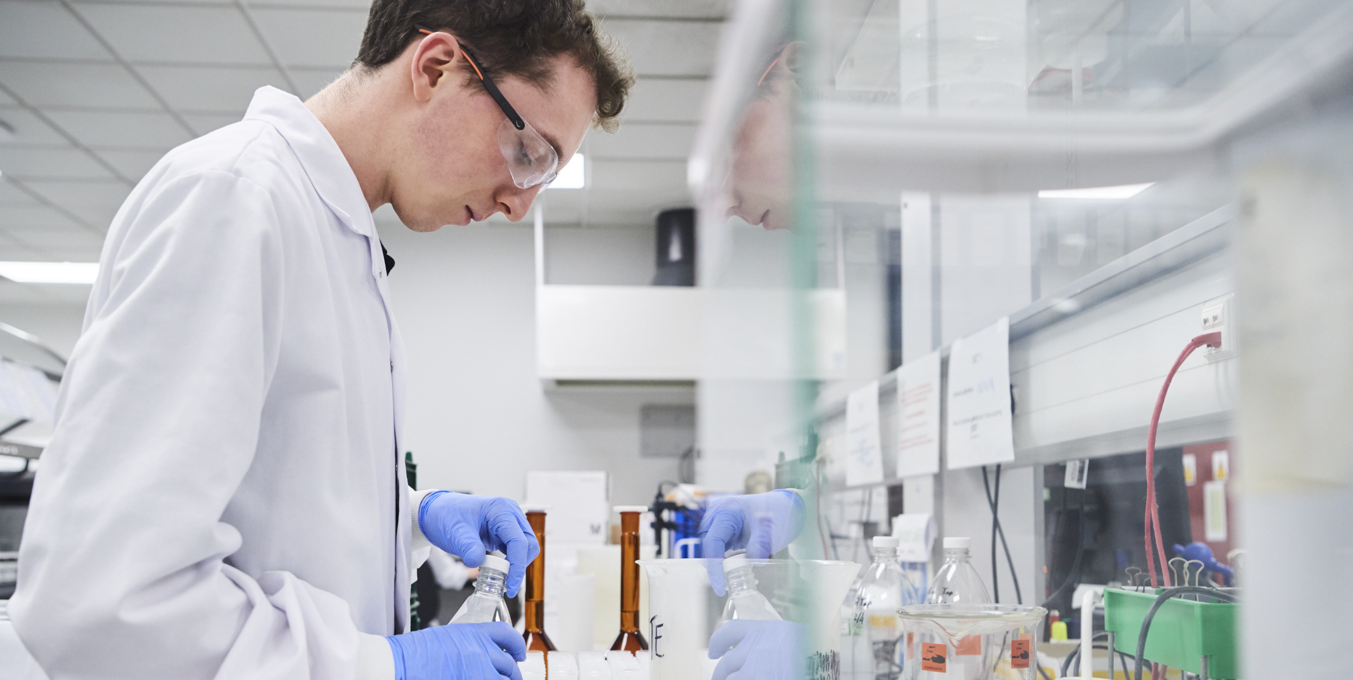 Male wearing goggles and white lab coat in lab environment unscrewing a cap on bottle