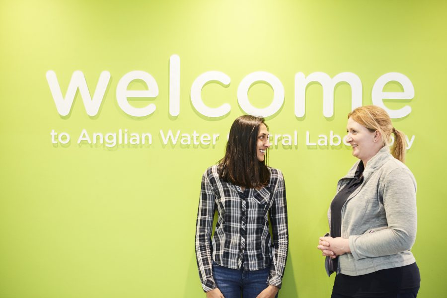 Two women smiling at each other in front of a wall with large letters on it reading Welcome to Anglian Water Central Laboratory